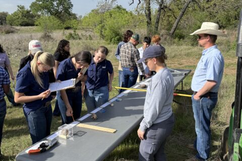 A group of people are gathered outdoors around a table with measuring equipment and papers. The scene is set in a grassy area with trees in the background. One person is standing at the end of the table, appearing to demonstrate something to the others who are observing intently.