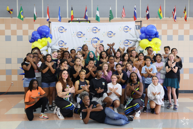 A group of people pose together indoors in front of a "STEM It Up" banner. They are standing on a checkered floor, surrounded by festive balloon arrangements. Flags of various countries hang from the ceiling, adding an international atmosphere.