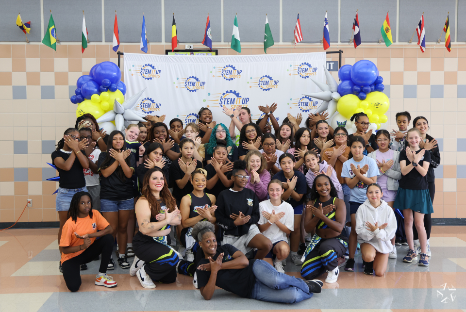 A group of people pose together indoors in front of a "STEM It Up" banner. They are standing on a checkered floor, surrounded by festive balloon arrangements. Flags of various countries hang from the ceiling, adding an international atmosphere.