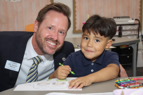 A smiling adult sits beside a child at a table in a classroom. The child is coloring with crayons on paper. A name tag on the adult reads "Dr. Steven Wurtz." Classroom supplies are visible in the background.