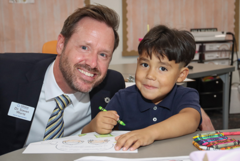 A smiling adult sits beside a child at a table in a classroom. The child is coloring with crayons on paper. A name tag on the adult reads "Dr. Steven Wurtz." Classroom supplies are visible in the background.