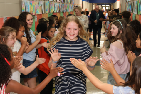 Students line a decorated school hallway, clapping and cheering as one student walks down the center, creating a celebratory tunnel.