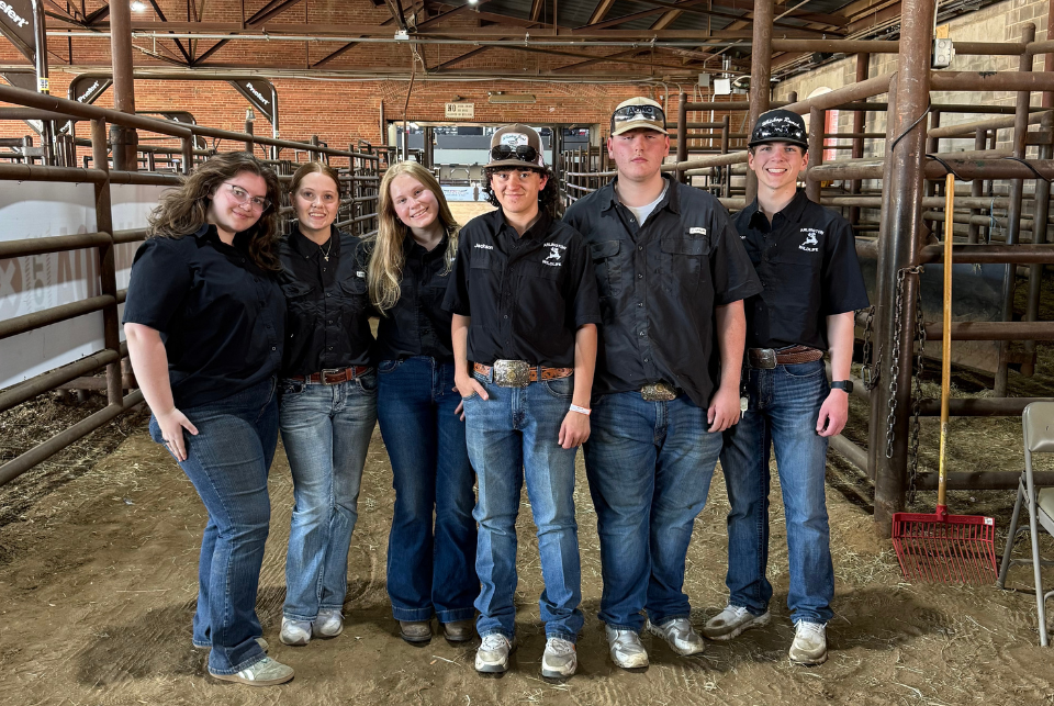 A group of six people stand side‑by‑side inside a livestock barn, all wearing dark shirts, jeans, and large belt buckles. Behind them are metal livestock pens, a brick wall, and a rake leaning against a chair. The setting appears to be an agricultural or livestock event, and the group poses together, smiling.