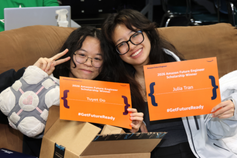 Two Martin High School seniors sit on a couch holding bright orange certificates announcing their 2026 Amazon Future Engineer Scholarship awards, smiling as they celebrate their achievement.