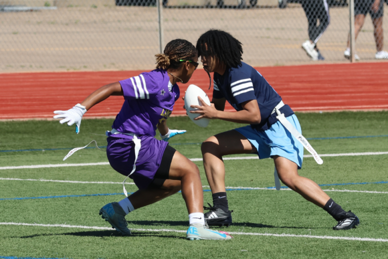 Two people play flag football on a green turf field. A player in a navy shirt and light blue shorts runs with a white football while a defender in a purple uniform reaches to grab their flag. Both wear flag belts with white flags. A red track, chain‑link fence, and a few spectators appear in the background.