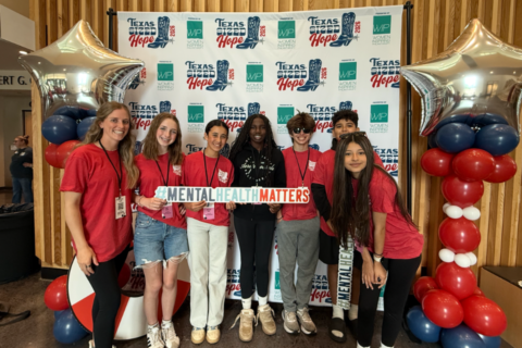 Students share hope during Hope Week. A group of seven people pose in front of a “Texas Sized Hope 2024 – Women Inspiring Philanthropy” backdrop. They stand between two balloon columns made of red, white, and blue balloons topped with silver star balloons. Most wear red shirts with name badges, while one person in the center wears a black hoodie. They hold a large sign that reads “#MENTALHEALTHMATTERS.