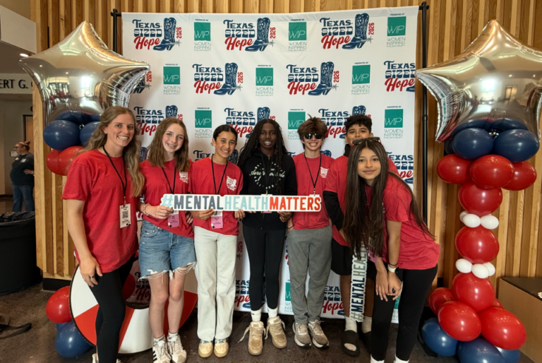 Students share hope during Hope Week. A group of seven people pose in front of a “Texas Sized Hope 2024 – Women Inspiring Philanthropy” backdrop. They stand between two balloon columns made of red, white, and blue balloons topped with silver star balloons. Most wear red shirts with name badges, while one person in the center wears a black hoodie. They hold a large sign that reads “#MENTALHEALTHMATTERS.