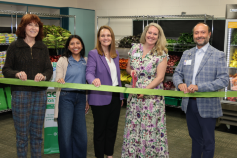 Five people stand in front of produce shelves during a ribbon‑cutting ceremony, holding a long green ribbon while the person in the center prepares to cut it with scissors. Fresh fruits and vegetables fill the shelves behind them, and a bilingual community‑focused sign is partially visible in the background.