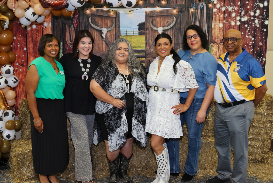 A group of six adults stands smiling in front of a western‑themed backdrop with barn doors, a cow skull decoration, string lights, hay bales, and brown‑white‑black cow‑print balloons. They are dressed in semi‑casual attire, with one person wearing cowboy boots. The setting resembles a country‑themed celebration.