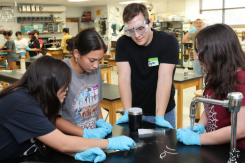 Students explore the future at quantum physics summer camp - A group of people in a science lab wearing protective gloves and goggles. They are gathered around a table observing an experiment with a container and some tools. Shelves with equipment are visible in the background.
