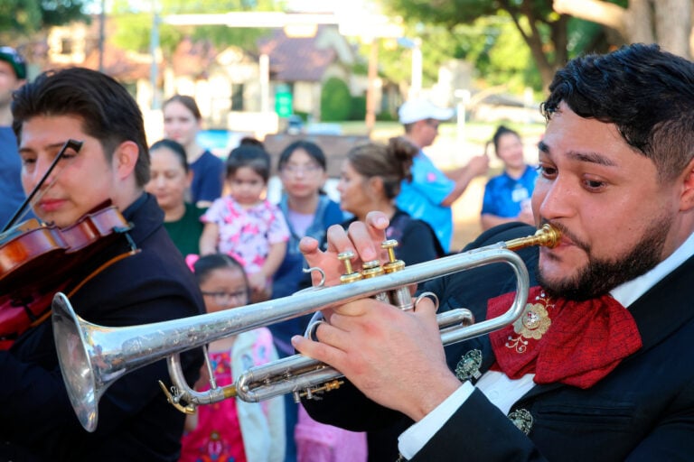 A musician in formal attire plays a trumpet while another plays a violin, with an audience of people watching in the background.