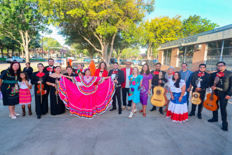 A group of people dressed in vibrant traditional attire, including musicians with instruments and a dancer in a colorful skirt, pose together outdoors in front of a school building.