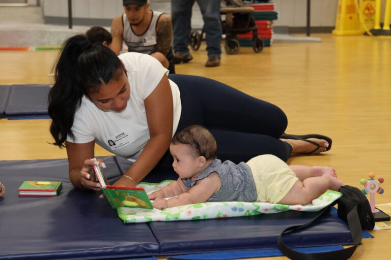 A person reads a book to a baby lying on a mat in a brightly lit indoor space.