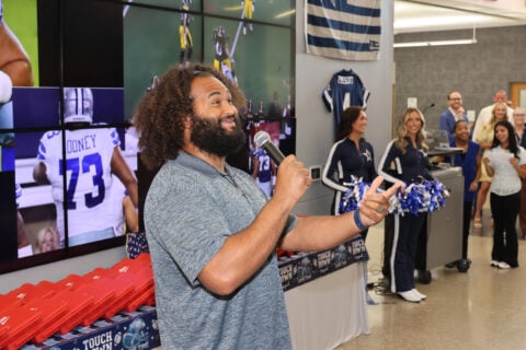 A person speaks into a microphone at an event, with Dallas Cowboys merchandise displayed around. Cheerleaders stand nearby, and a wall of TV screens shows a football game in the background.