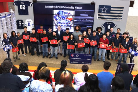 A group of people is standing on a stage holding certificates, with a display screen behind them showing "Automotive S2000 Scholarship Winners" and logos of Ford, the NFL, and the Dallas Cowboys. Dallas Cowboys jerseys and a large check are also visible. An audience is seated in the foreground.