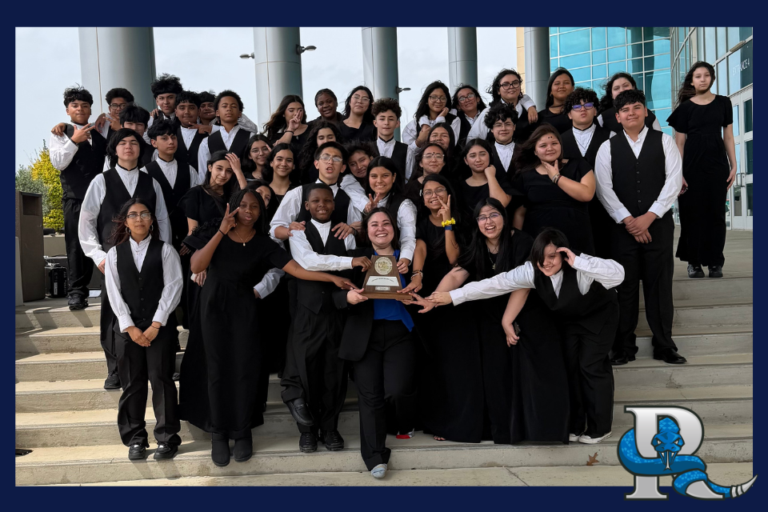 A large group of students in formal black concert attire stand on outdoor steps in front of a modern building. They pose together around a central student holding a trophy, celebrating a performance or competition win. A blue serpent‑wrapped “P” logo appears in the bottom right corner.