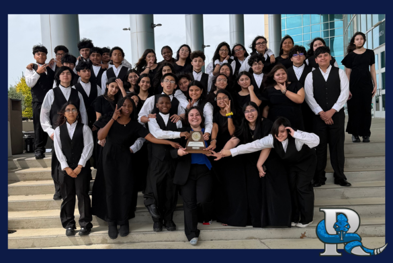 A large group of students in formal black concert attire stand on outdoor steps in front of a modern building. They pose together around a central student holding a trophy, celebrating a performance or competition win. A blue serpent‑wrapped “P” logo appears in the bottom right corner.