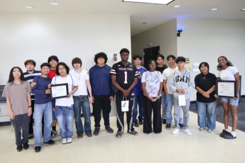 A diverse group of people stands together in a well-lit indoor space. One person in the center is wearing a Texas Tech jersey, while two others hold certificates. There are signs in the background indicating room numbers.