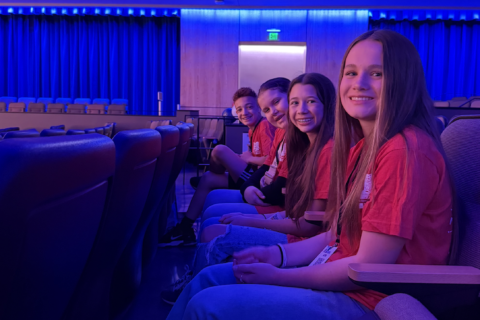 Group of students sitting in a row in the auditorium. They are wearing red shirts and smiling.