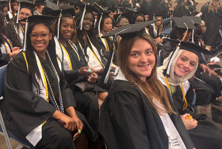 A group of people in graduation caps and gowns are seated in rows, smiling. Some are wearing honor cords or stoles, and they are inside a large venue. The atmosphere is celebratory.